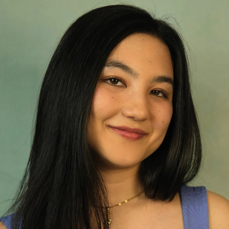 Sara Ross Avery, a woman with long, straight black hair and light skin, smiles softly. She wears a light purple sleeveless top and a delicate gold necklace. The background is plain and softly blurred in shades of green and beige.