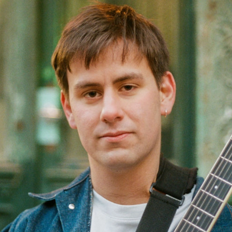 Miles Mindlin, a young person with short brown hair and a slight smile, gazes at the camera while wearing a blue jacket over a white shirt and an earring. They hold a guitar, with a blurred green background behind them.