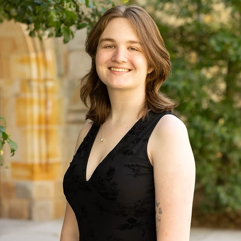 A young woman with shoulder-length brown hair smiles outdoors. Isabelle Millman wears a sleeveless black dress and a small necklace. Green trees and a stone archway are in the blurred background, suggesting a garden or campus setting.