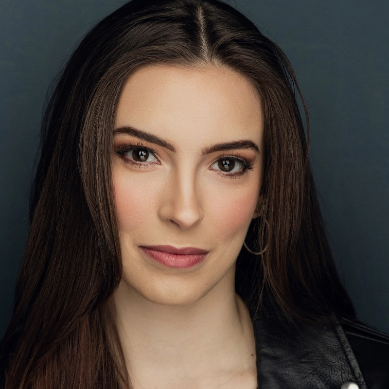 A young woman with long brown hair and brown eyes looks directly at the camera. She has light skin, natural makeup, and wears a black leather jacket. The background is a solid dark color, giving the portrait a professional and polished look.