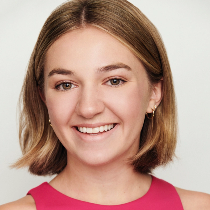 A young woman with straight, shoulder-length light brown hair smiles at the camera. She wears a sleeveless pink top and small gold hoop earrings. The background is plain and light-colored, and the lighting is bright and even.