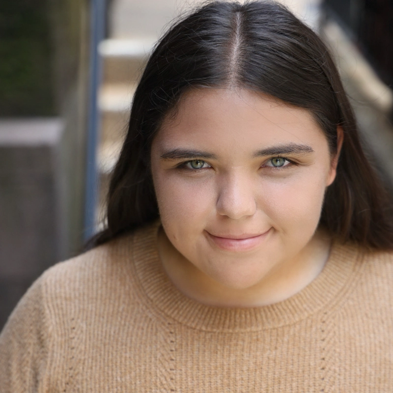 A young person with long, straight brown hair and light eyes smiles gently at the camera. They wear a tan knit sweater and stand outside on steps, with soft natural light illuminating their face. The background is blurred.