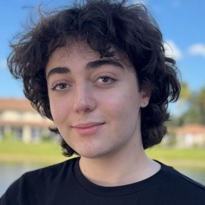 A young person with fair skin, wavy brown hair, and dark eyes smiles softly at the camera. They are wearing a black shirt. In the blurred background, houses and green grass hint at a calm day by the water under Alexander Mozes’s blue sky.