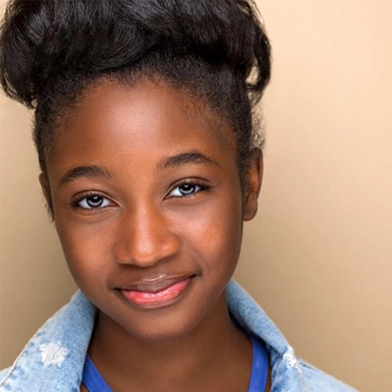 Nadia Daniel, a young Black girl with smooth skin and natural hair styled in a high bun, smiles gently. She wears a light blue denim jacket over a blue shirt, posing confidently against a plain beige background.