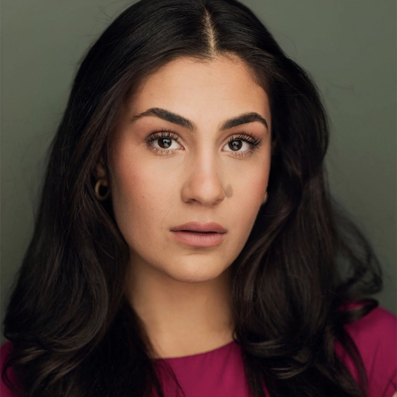 A woman with long, dark wavy hair and strong eyebrows looks directly at the camera. Andrea Nalbandian wears warm-toned makeup, a magenta top, and has a beauty mark on her left cheek. The plain background draws focus to her face.