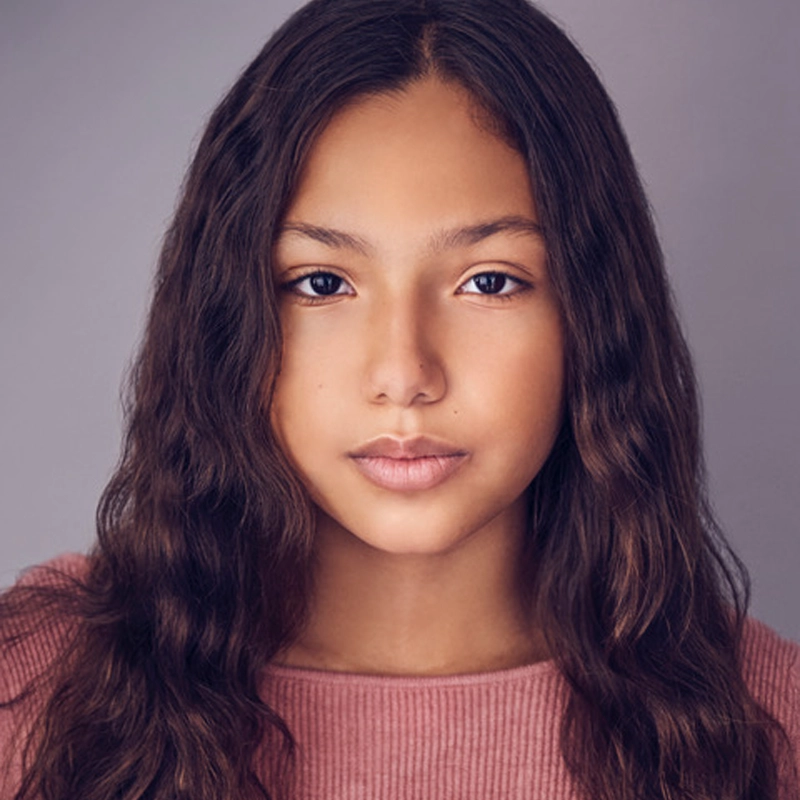 A young girl with long, wavy brown hair and brown eyes looks directly at the camera. Angeli Negron is wearing a pink ribbed top and has a neutral expression, set against a soft, blurred gray background that draws focus to her face.