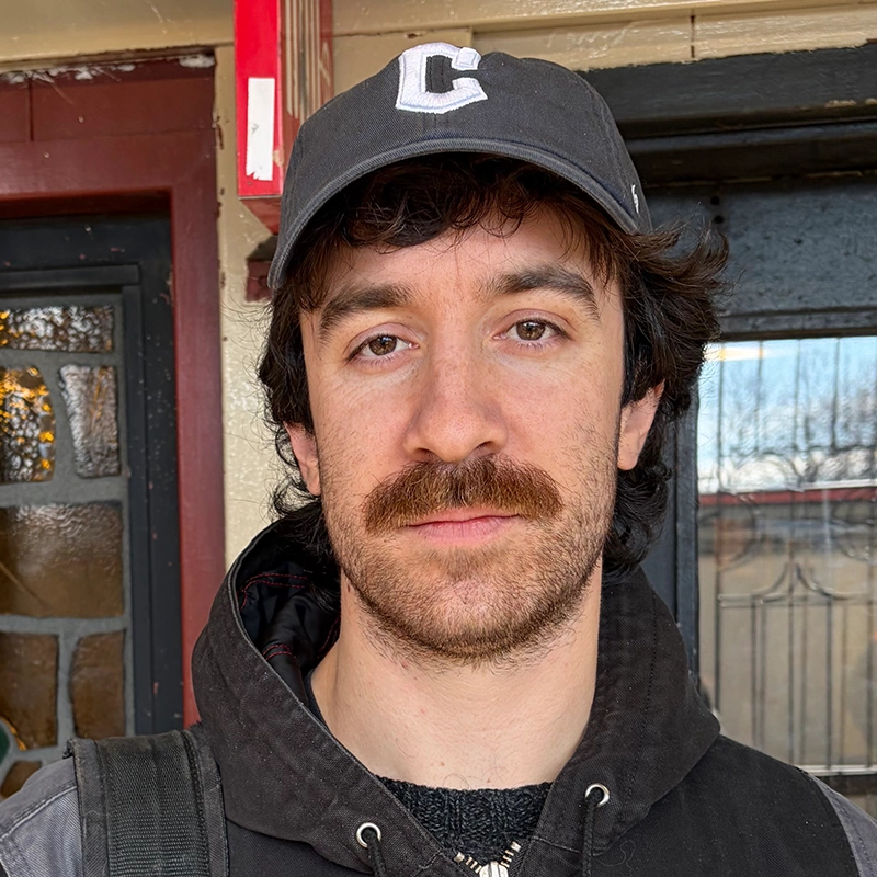 A man with wavy brown hair, a full mustache, and light skin wears a black baseball cap with a white “C,” a black jacket, and a backpack strap. He stands in front of Hannah Mayer’s house with wooden trim and a glass panel door.