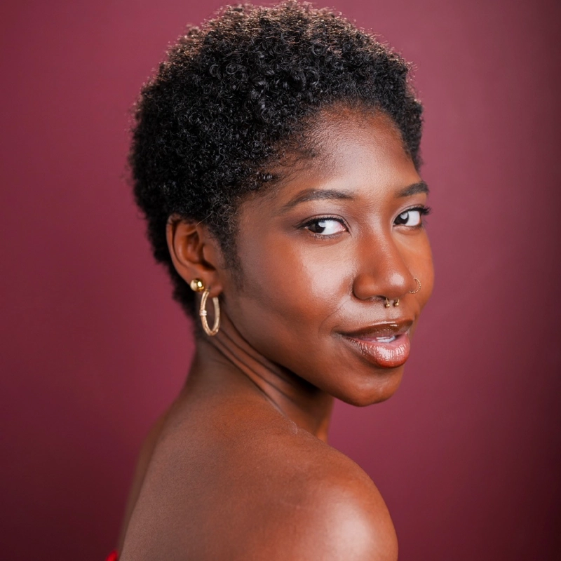 A woman with short, curly black hair poses in front of a maroon background. She looks over her shoulder, smiling slightly. She wears gold hoop earrings, a septum ring, and a strapless top, with smooth, glowing skin and natural makeup.