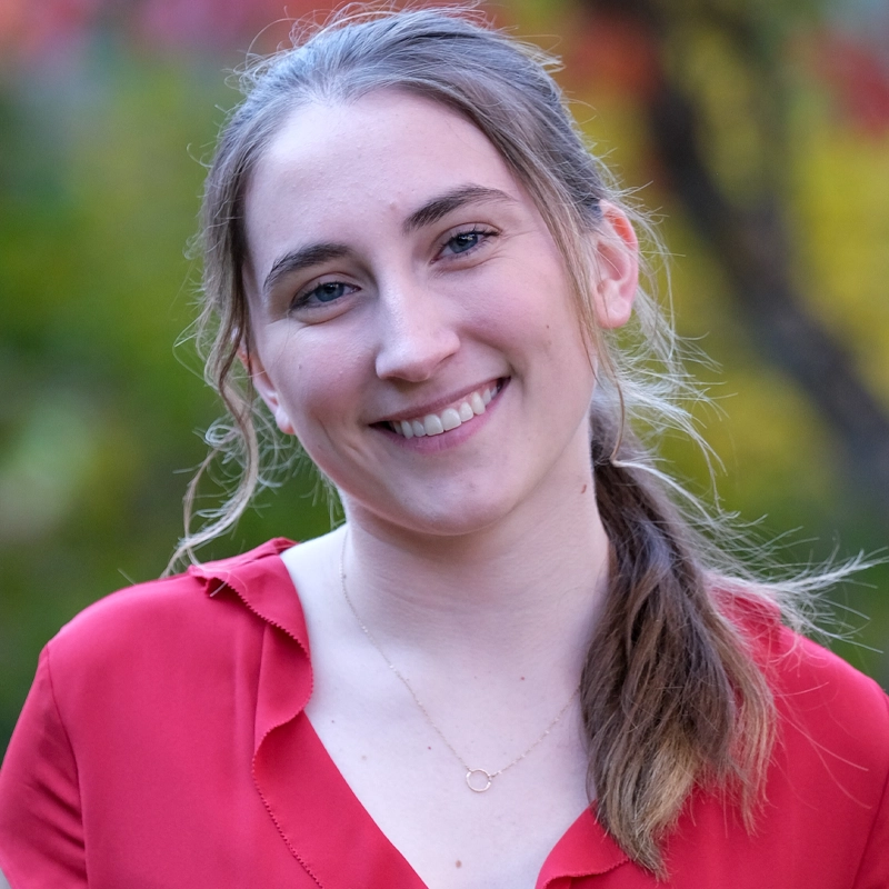 A young woman with light skin and long brown hair tied in a ponytail smiles at the camera. She wears a red blouse and a delicate gold necklace. The background is blurred with green and hints of autumn colors.
