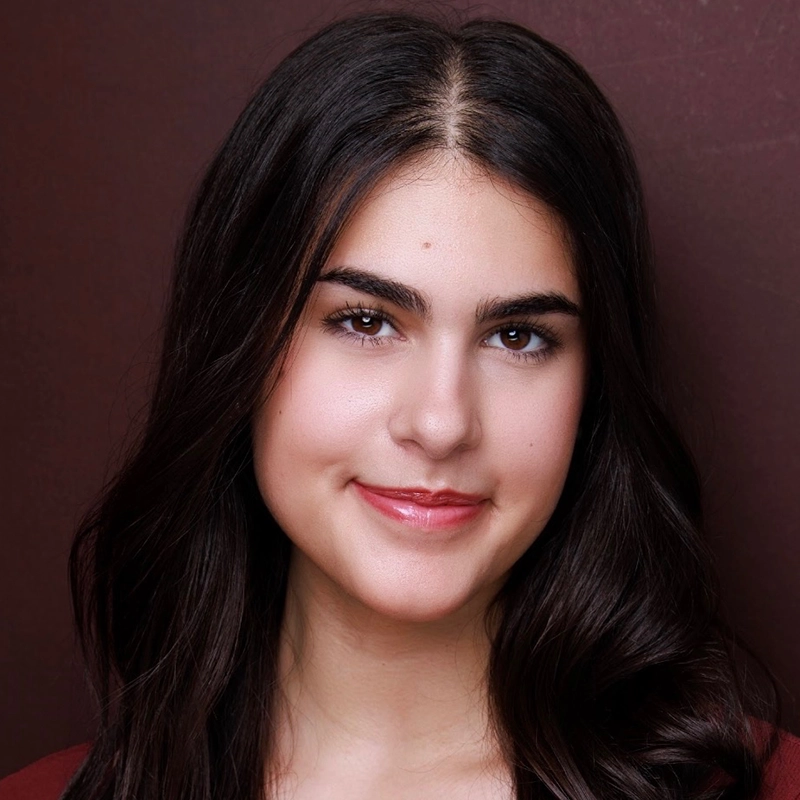 A young woman with long, dark brown hair and fair skin smiles gently. She has well-groomed eyebrows, brown eyes, and wears natural makeup with pink lip gloss. The background is plain and dark burgundy, highlighting her face.