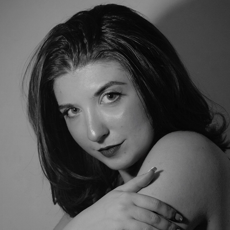 A young woman with shoulder-length dark hair looks directly at the camera. Lexis Trechak has light skin, dark lipstick, and subtle makeup. Her right arm crosses over her bare shoulder as she poses in black and white with soft, dramatic lighting.