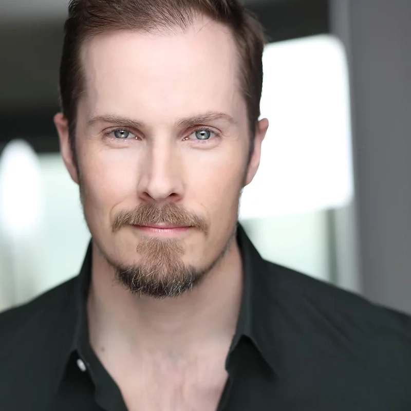 Michael Padgett, a man with fair skin, light blue eyes, and short brown hair, has a trimmed beard and mustache. He wears a dark green collared shirt and looks confidently at the camera, with soft natural light in the blurred background.