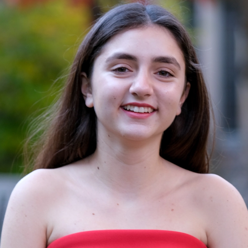 Isabel Baxter, a young woman with long brown hair, smiles outdoors in Paris. She is wearing a red, off-the-shoulder top. The background is blurred with green foliage and bright natural light, evoking a charming outdoor setting.
