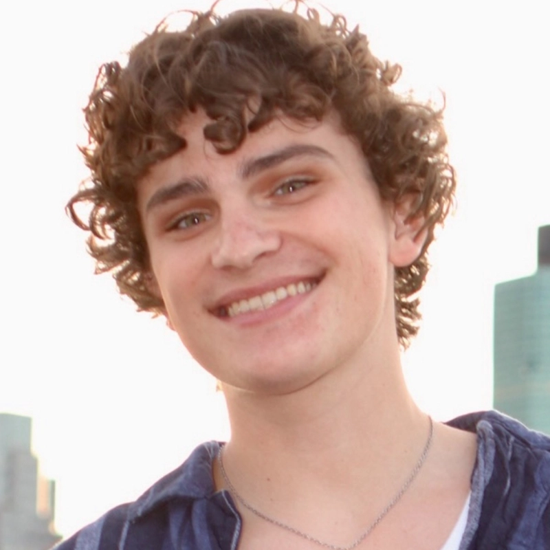A young person with curly brown hair and light skin smiles at the camera. Weston Paskewich wears a blue shirt over a white top and a silver necklace. The background features an out-of-focus city skyline with tall buildings and bright sky.
