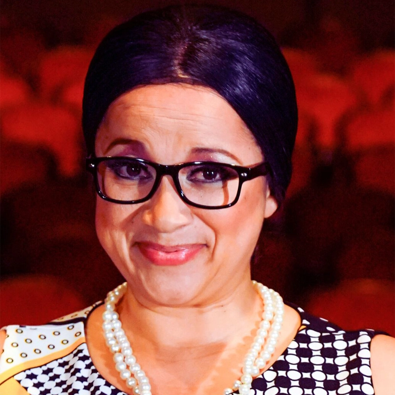 Martha Payares, with dark hair in an updo, wears black glasses, a black-and-white patterned dress, and a pearl necklace. She smiles gently as the theater’s red seats blur in the background, warm lighting highlighting her face.