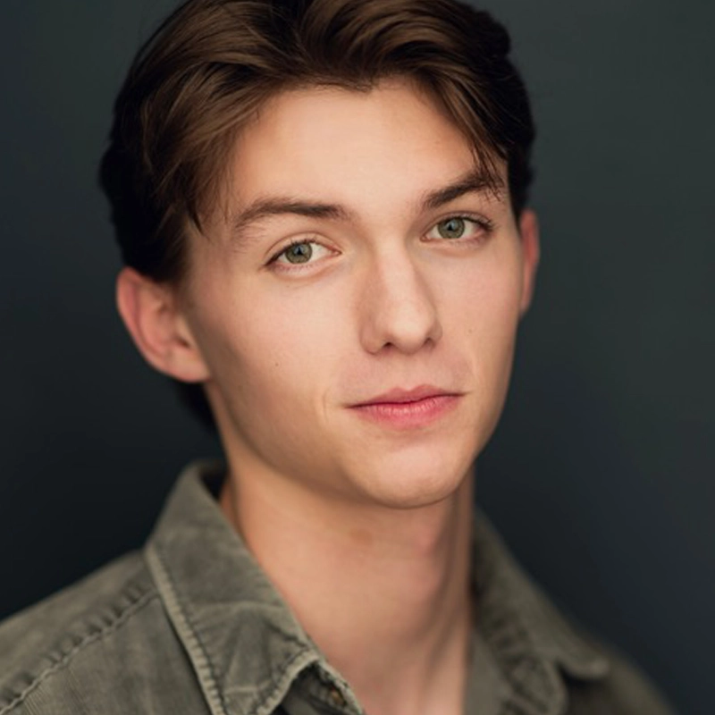 A young person with fair skin, short brown hair, and green eyes looks at the camera with a neutral expression. They are wearing a grayish-green button-up shirt over a dark background. The lighting is soft, highlighting their facial features.