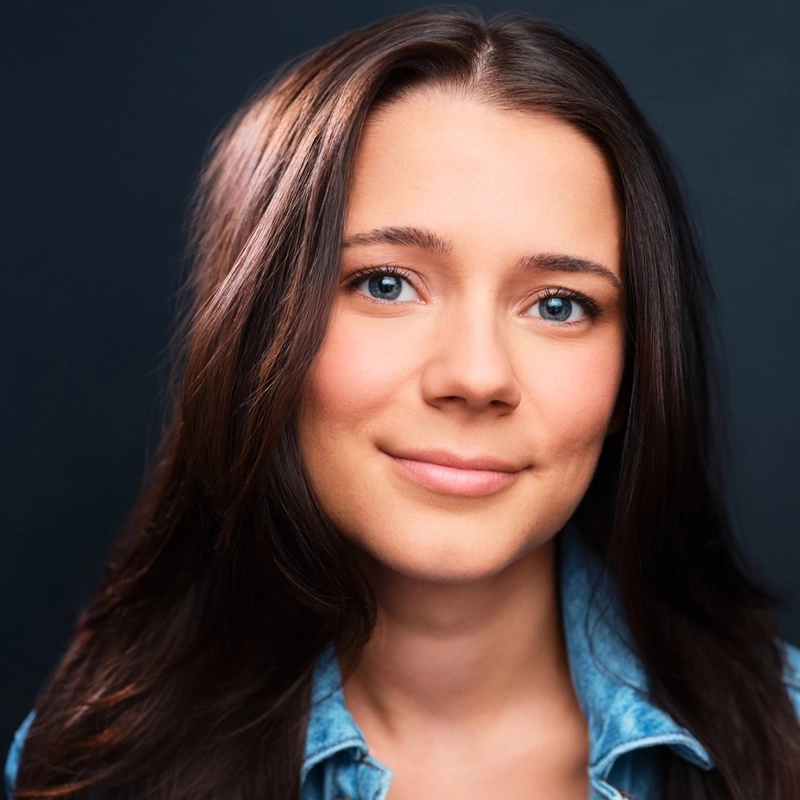 Ashley Poulin, a young woman with fair skin, long brown hair, and blue eyes, smiles gently. She wears a light blue denim shirt and is posed against a dark background, with soft, even lighting on her face.