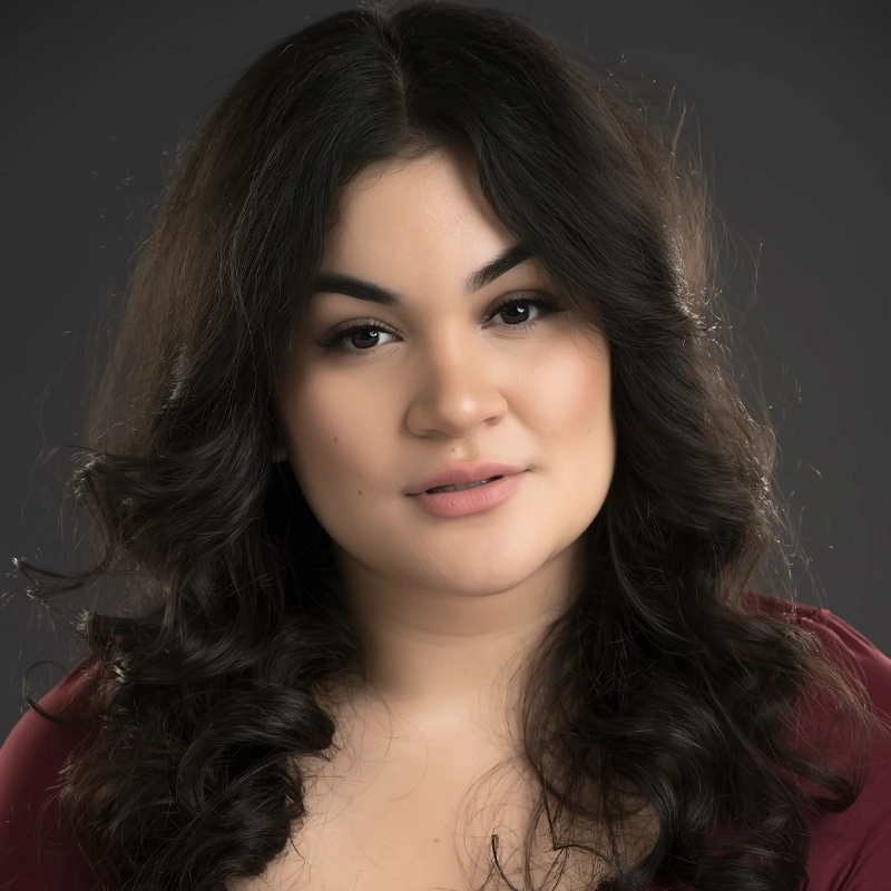 Julianna Ramos, a young woman with long, dark curly hair and defined eyebrows, poses against a dark, blurred background. She has light skin, natural makeup, and wears a maroon top, looking confidently into the camera with a neutral expression.