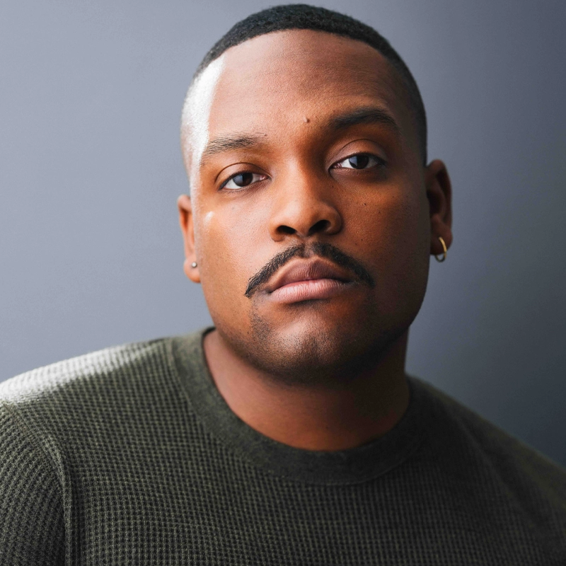 A man with short hair, a trimmed mustache, and small hoop earrings wears a textured dark green sweater. He looks directly at the camera with a neutral expression against a plain gray background. Soft lighting highlights his face.