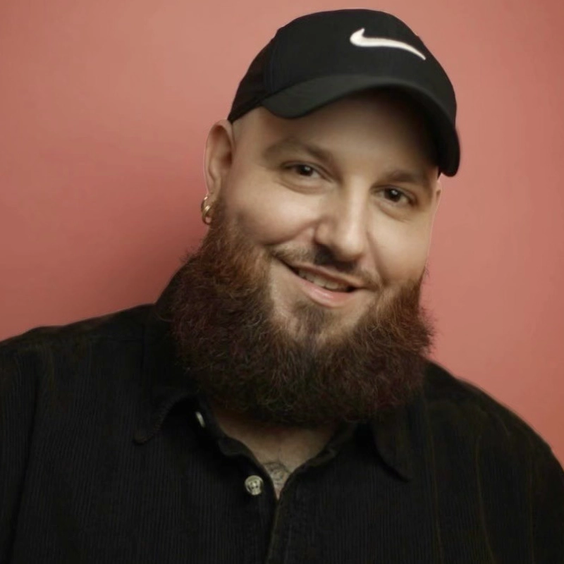 A bearded man with light skin smiles at the camera. Justin Reamy wears a black Nike cap, a black button-up shirt, and gold hoop earrings. The background is a solid, muted pink color.