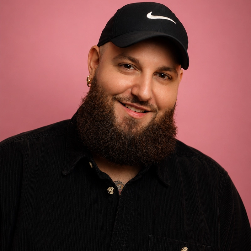 A bearded man, Justin D. Reamy, with a black Nike cap, black button-up shirt, and gold hoop earrings smiles against a pink background. He has a friendly expression and neatly groomed beard. He faces the camera, showing confidence and approachability.