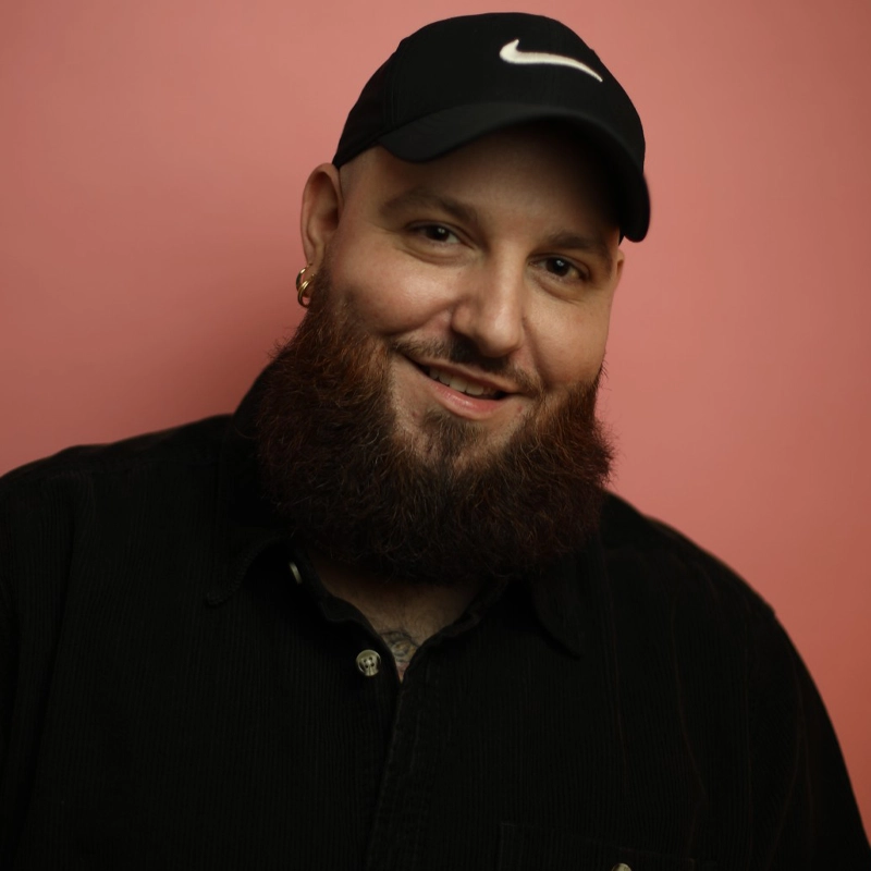 A bearded man, Justin Reamy, wearing a black cap with a white Nike logo, gold hoop earrings, and a black button-up shirt smiles at the camera. He stands against a plain pink background.