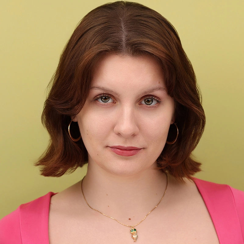 A young woman with short brown hair and hoop earrings looks at the camera against a light green background. She wears a pink top and a gold necklace with an owl pendant, and has a calm, confident expression.