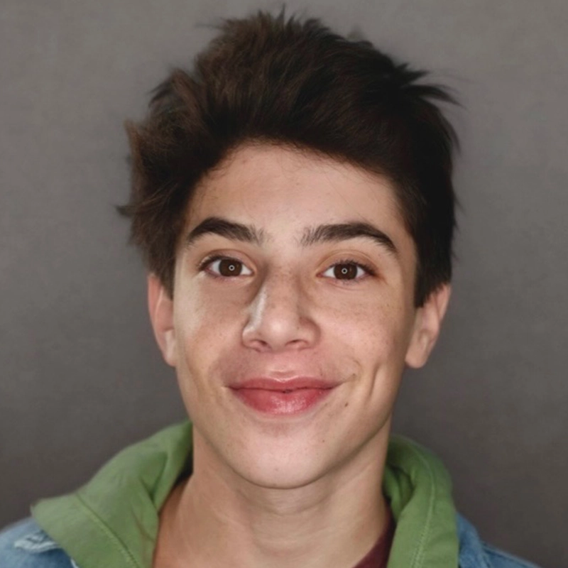 Micah Wolfensohn, a young person with short, dark, tousled hair, smiles slightly at the camera. They have brown eyes, dimples, and light skin with freckles. They're in a green hoodie under a blue jacket against a plain gray background.
