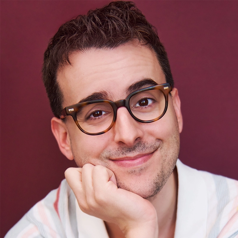 Christina Acosta Robinson, with short dark hair, light skin, and brown-framed glasses, smiles gently at the camera. Resting her chin on her hand and wearing a light shirt, she poses before a solid, deep burgundy background.