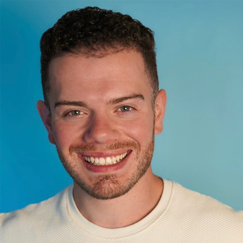 A young man with short, curly brown hair and trimmed beard smiles widely. He wears a white textured shirt and stands in front of a bright blue background, looking directly at the camera with a cheerful expression.
