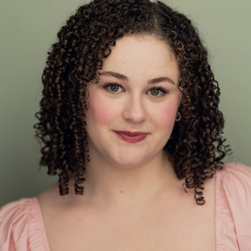 A woman with fair skin and tightly curled brown hair smiles softly. Reminiscent of Daphne Riddle, she wears natural makeup with rosy cheeks and pink lipstick, dressed in a light pink top with puffed sleeves against a muted green background.