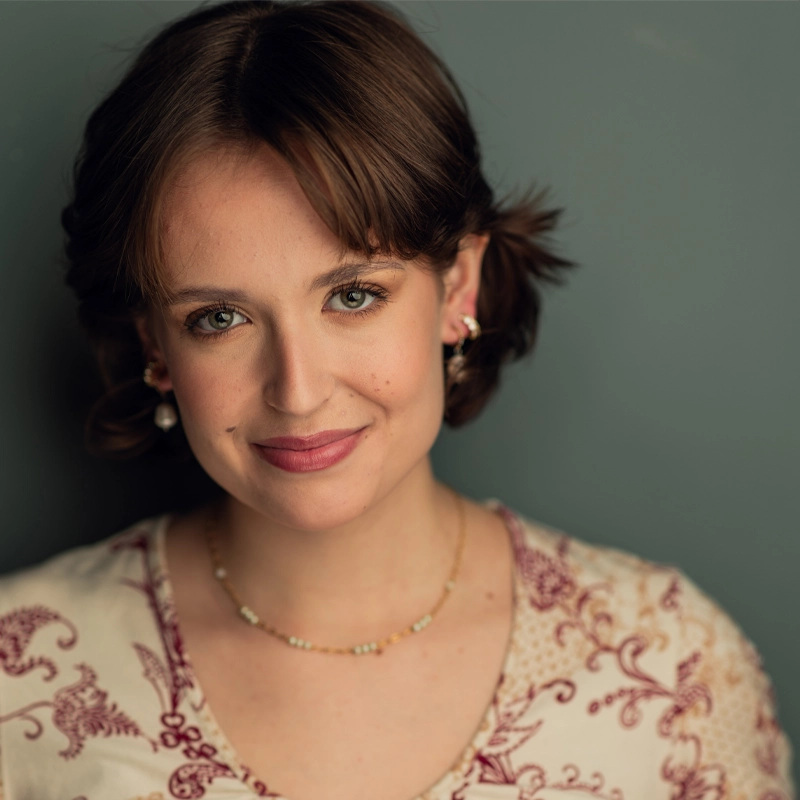 A young woman with short brown hair styled in loose waves smiles gently. She wears pearl earrings, a delicate beaded necklace, and a cream blouse with red floral patterns. The background is plain and muted green, giving a soft, warm atmosphere.