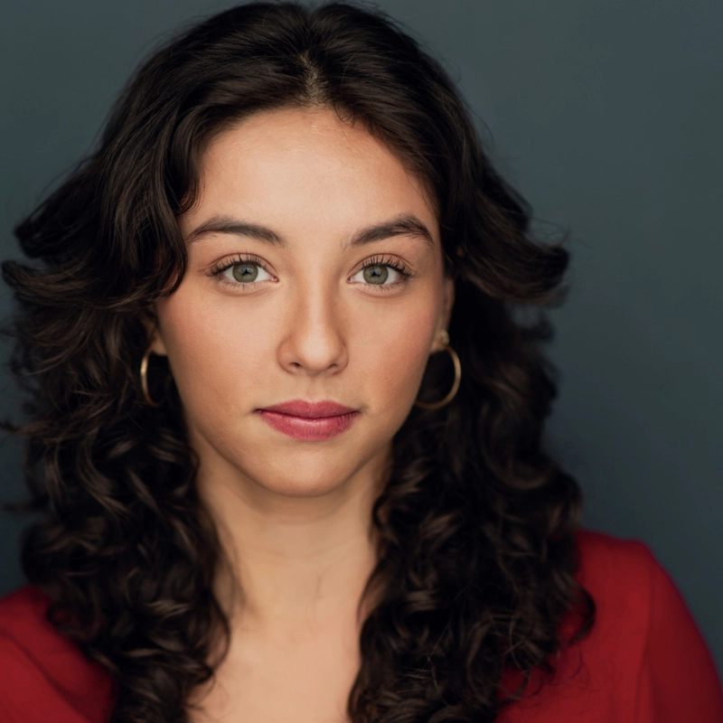 Andrea Paulina Robles, a young woman with long, dark curly hair and green eyes, looks directly at the camera. She wears gold hoop earrings and a red top against a plain, dark blue-gray background, her expression neutral.