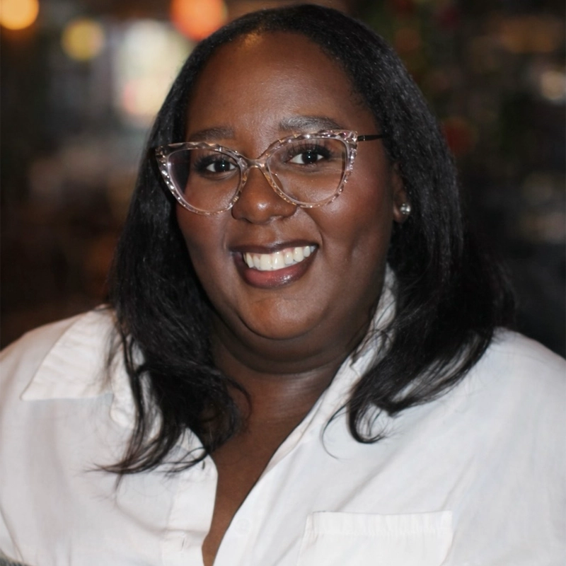 A woman with dark skin and straight black hair smiles warmly. She wears clear, stylish eyeglasses and a white collared shirt. The background is softly blurred with warm lighting, creating a cozy and inviting atmosphere.