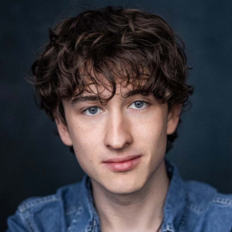 Lexis Trechak, a young person with fair skin, curly brown hair, and blue eyes, looks directly at the camera with a slight smile. Wearing a blue denim shirt against a dark, blurred background, even lighting highlights their facial features.