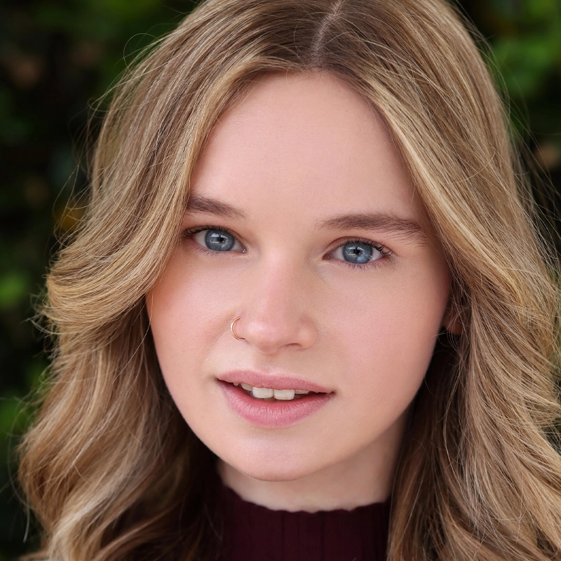 A young woman with wavy, light brown hair and blue eyes looks at the camera. She has light skin, a small nose ring, and wears a burgundy top. The background is blurred greenery, drawing focus to her face.