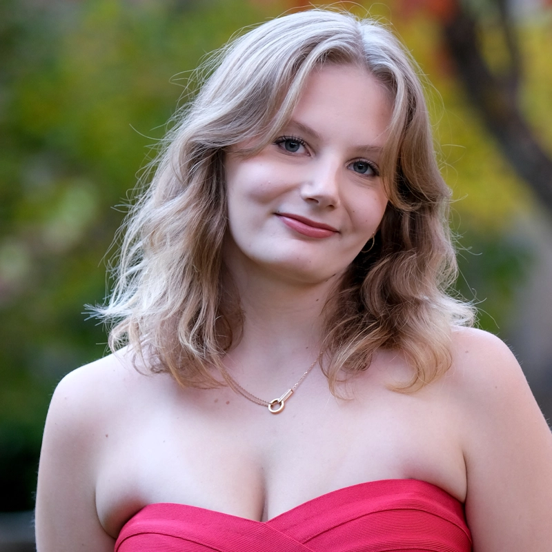 A young woman with wavy blonde hair and fair skin smiles gently at the camera. She is wearing a strapless pink dress and a gold necklace with a small pendant. The background is blurred with green and yellow foliage, suggesting an outdoor setting.