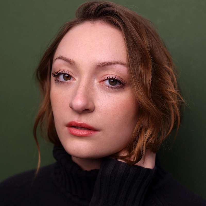 A young woman, Elena Salzberg, with wavy light brown hair parted to the side, wearing a black turtleneck sweater, poses against a green background. She gazes calmly at the camera, her hand touching her neck, with natural makeup and subtle pink lips.