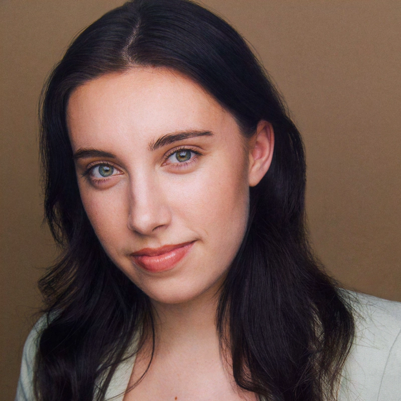 A young woman with fair skin, long dark brown hair, and green eyes gazes softly at the camera. She is wearing a light-colored top and is posed against a plain beige background, giving the portrait a warm, natural feel.