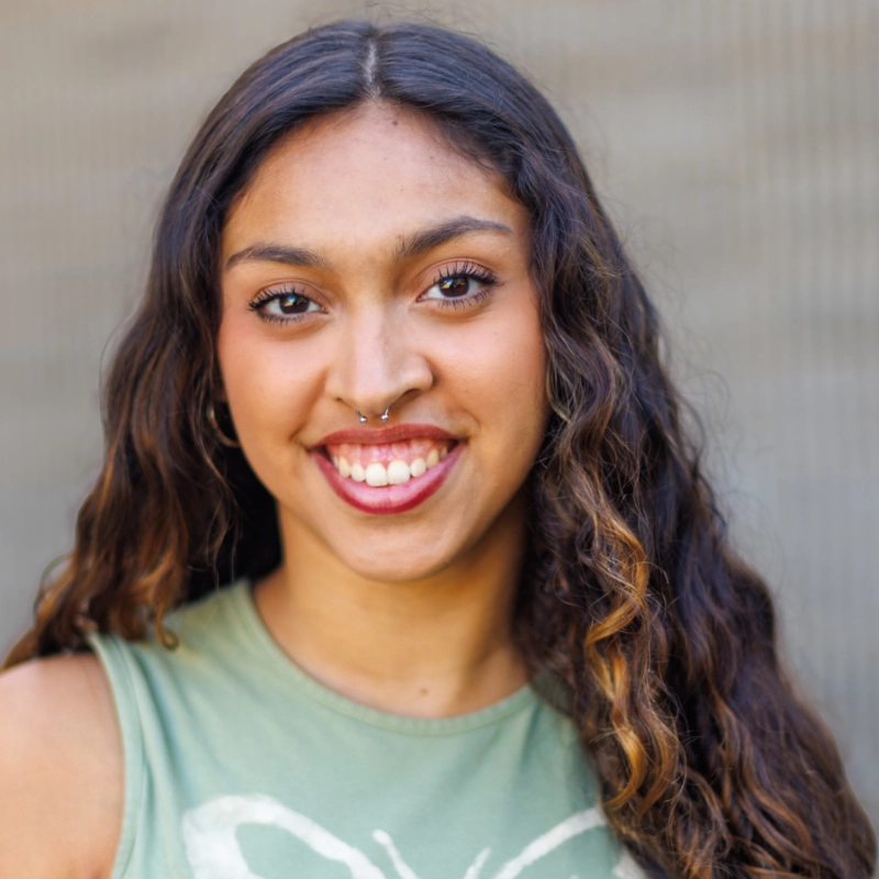 Chelsea Santiago, a young woman with long, wavy brown hair and medium skin, smiles warmly at the camera. She has a septum piercing and wears a sleeveless green top with a white design against a softly blurred neutral background.
