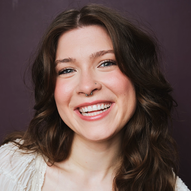 Sara Ross Avery, a young woman with wavy brown hair, smiles broadly, showing her teeth. She has a nose ring, light skin, blue eyes, and wears natural makeup. Dressed in a cream-colored top, Sara stands in front of a solid dark purple background.