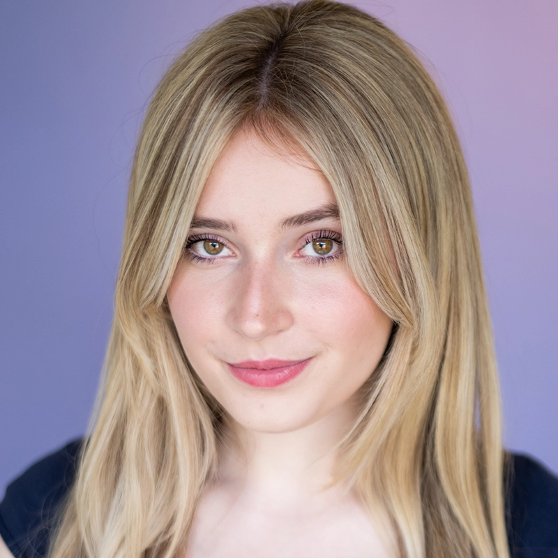 Addison Zacharias, a young woman with long, straight blonde hair and green eyes, looks at the camera. She has light skin, subtle makeup, and a slight smile. Her dark top contrasts with the smooth purple and blue gradient background.