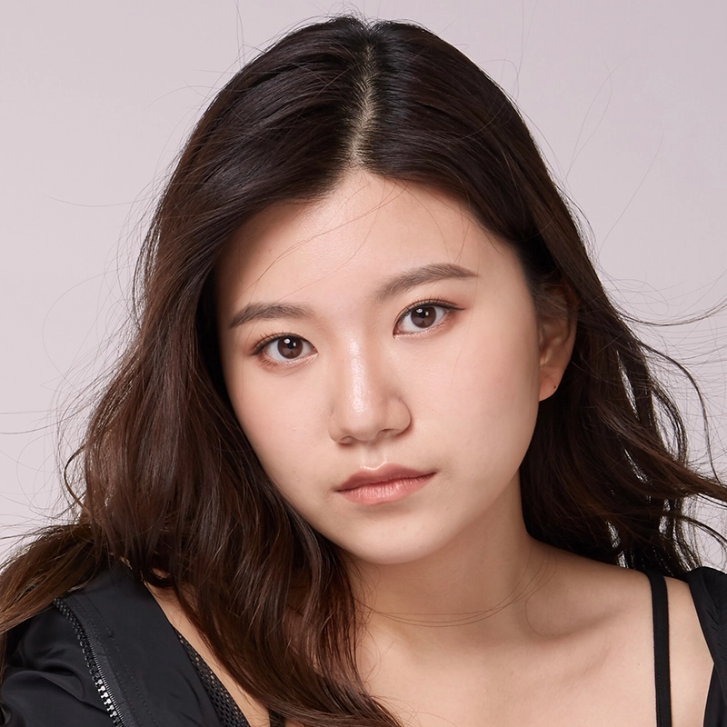 A young woman, Jenna Willet, with long, wavy dark hair looks directly at the camera with a neutral expression. She has light skin, natural makeup, and wears a black top against a plain light background. Strands of hair fall gently around her face.