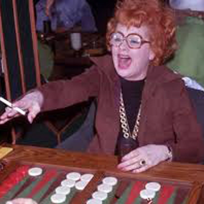 Woman with curly red hair, glasses, and a brown outfit, holding a cigarette and a Vodka Stinger, excitedly playing backgammon. She reaches out with her left hand, mouth open in laughter or shout, seated at a table with red and white game pieces.