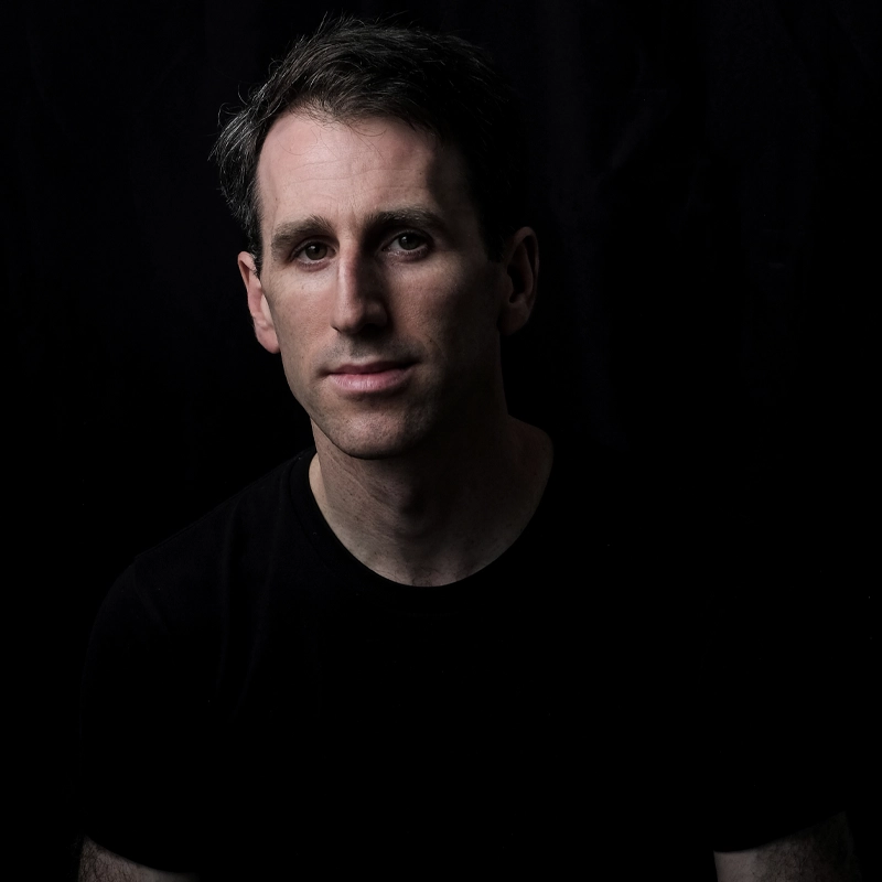 A man with short dark hair and some gray stands against a black background, wearing a plain black shirt. JP Schlegelmilch looks directly at the camera, half of his face softly illuminated while the rest blends into the shadows.