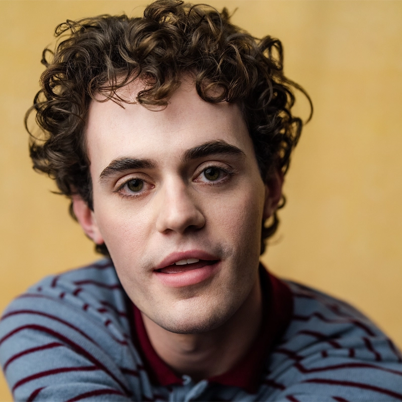 A young person with fair skin, short curly brown hair, and brown eyes is gazing directly at the camera. Noah Schuster wears a blue sweater with maroon stripes and a maroon collar. The soft yellow background enhances their relaxed, friendly expression.