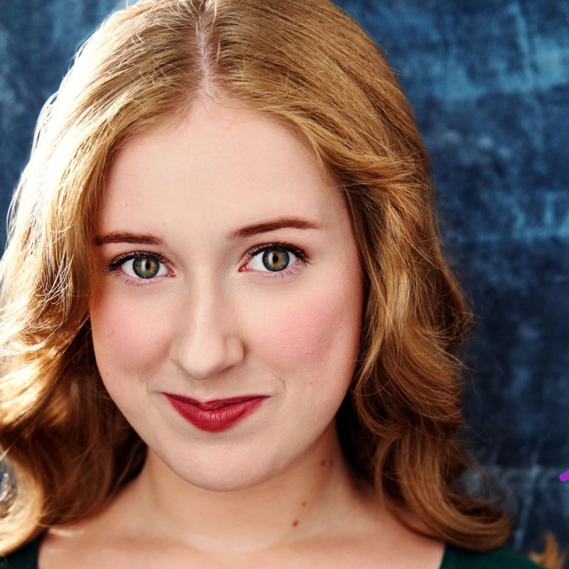 Clare Shannon, a young woman with fair skin and wavy, light brown hair, smiles gently. She has green eyes, rosy cheeks, and wears subtle makeup with red lipstick. Dressed in a dark green top, she stands against a mottled blue background.