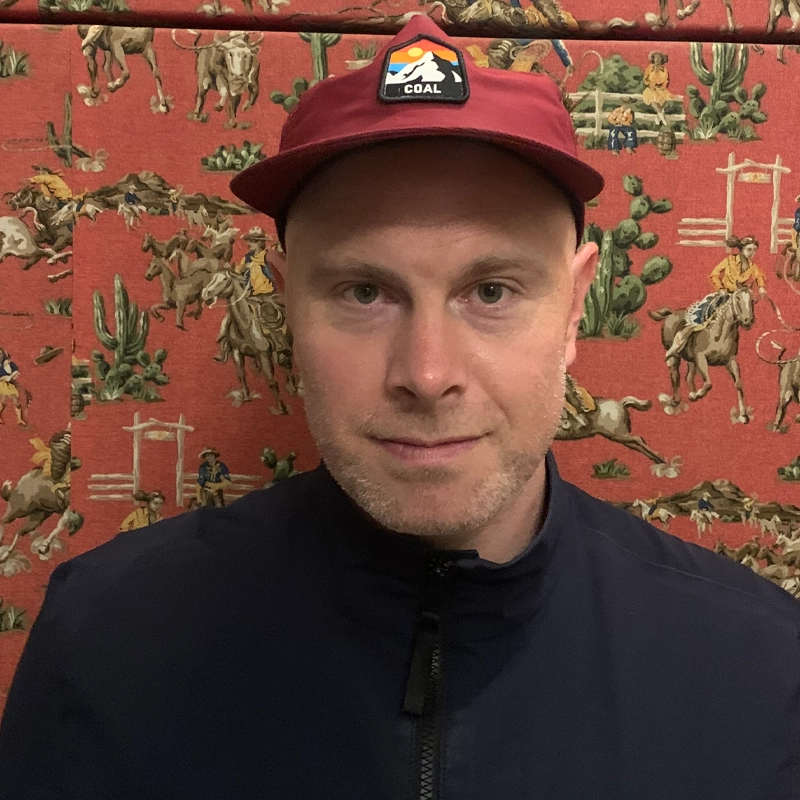 A man in a red cap with a mountain patch faces the camera, wearing a dark jacket. The background is red wallpaper featuring illustrations of cowboys riding horses and cowboy-themed scenery with cacti and gates.