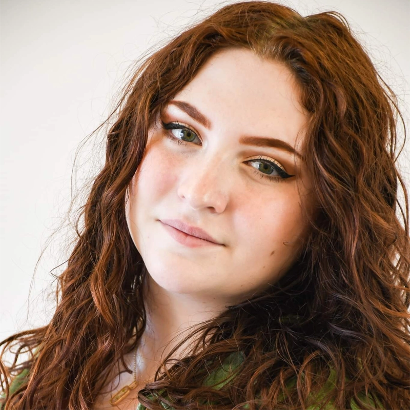 A young woman with long, wavy auburn hair and fair skin looks at the camera with a gentle smile. Gretchen Shope has green eyes, prominent eyebrows, subtle makeup, and wears a green top against a plain white background.