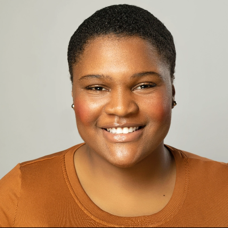 Aniya Simmons, with short curly hair, smiles warmly. She wears stud earrings and an orange-brown textured top. The plain light gray background highlights her friendly expression and glowing complexion.
